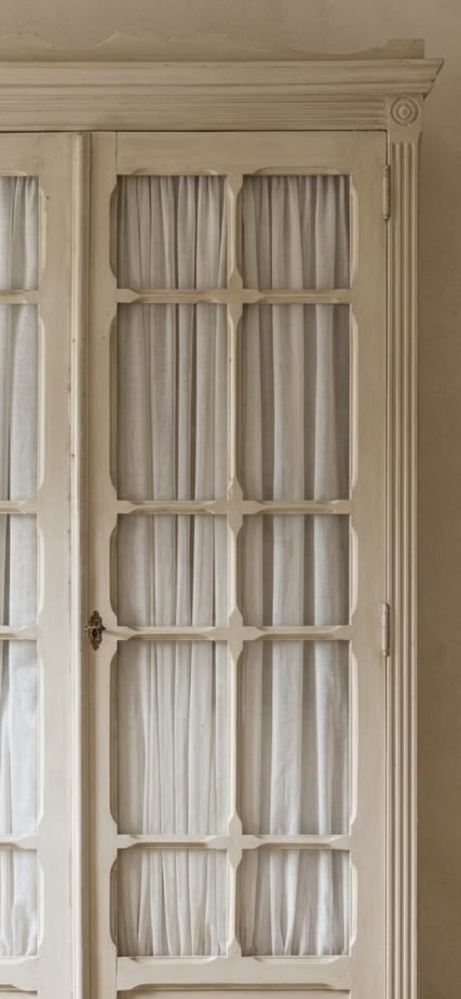 A painted glazed cabinet with a carved cornice and gathered linen behind the glass.