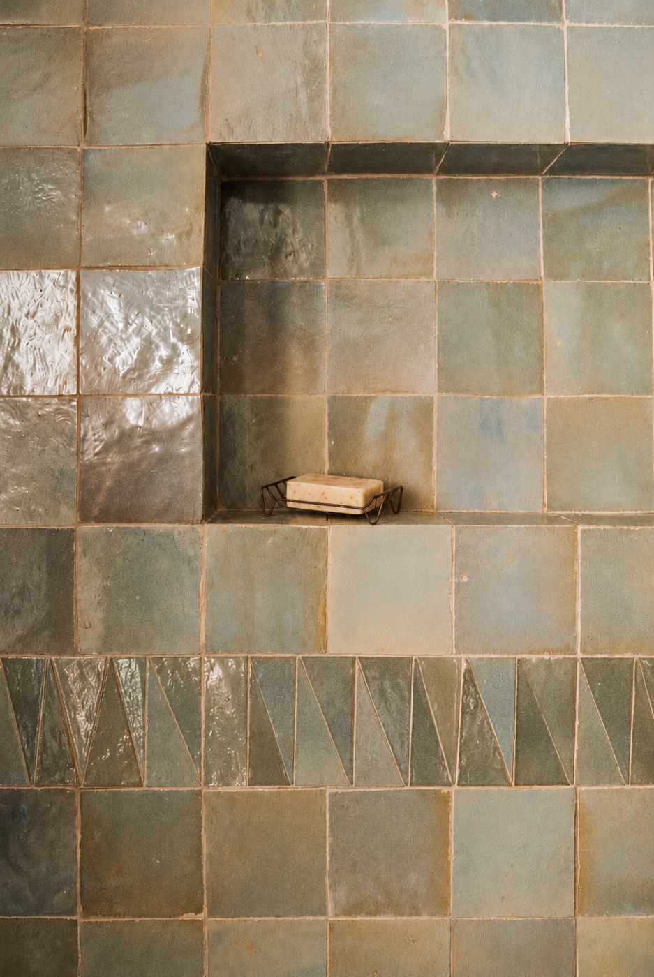 Handmade sage zellige tiles around a shower niche, with a band of triangle tiles and a bar of soap on a wire dish.